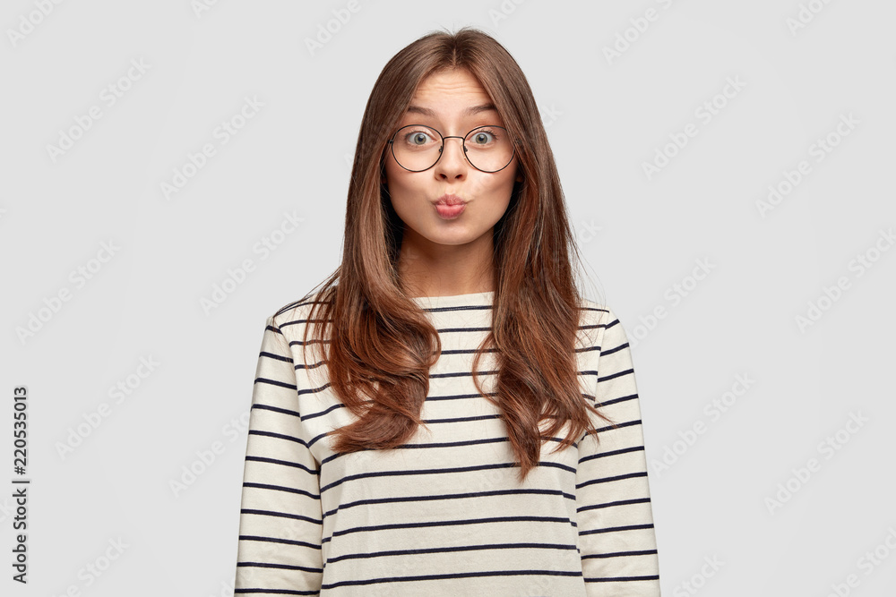 Lovely attractive woman pouts lips at camera, going to kiss someone on distance, has pleasant appearance, dressed in casual clothes, poses against white background. Human facial expressions. Lovely attractive woman pouts lips at camera, going to kiss someone on distance, has pleasant appearance, dressed in casual clothes, poses against white background. Human facial expressions.