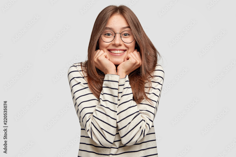 Indoor shot of happy beautiful student keeps hands under chin, glad to recieve praise, wears round spectacles, spends free time with close friends, stands against white background. Happiness concept Indoor shot of happy beautiful student keeps hands under chin, glad to recieve praise, wears round spectacles, spends free time with close friends, stands against white background. Happiness concept