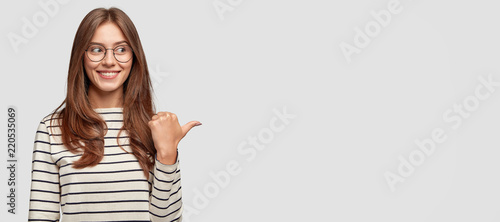Indoor shot of pretty young brunette woman with dark hair, notices funny scene left aside, points with thumb, being in good mood, has joyful and carefree expression, isolated over white background photo