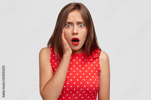 Reaction and emotions concept. Shocked emotional gorgeous young female person exclaims with stupefaction, hears bad negative news, wears red polka dot dress, stands against white background.