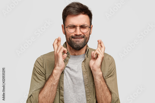 Portrait of handsome unshaven guy presses lips, closes eyes, keeps fingers crossed, has strong believe in good luck before important trip, wills everything would be fine. God, help me please!