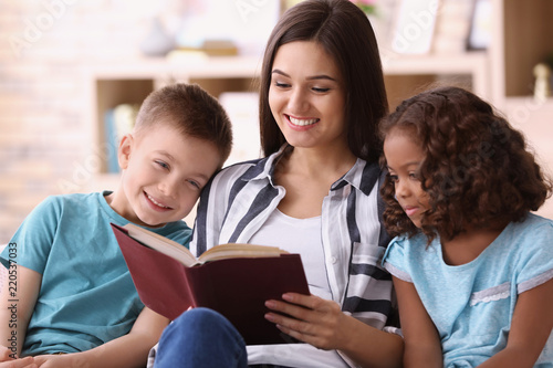 Young woman reading book to little kids indoors. Child adoption