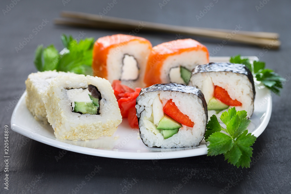 Sushi on a square white plate with parsley and chopsticks on a dark background. Sushi on a square white plate with parsley and chopsticks on a dark background.
