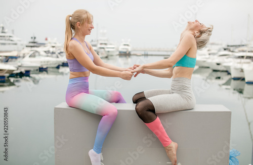 Two caucasian girlfriends wearing sportive outfit relaxing after outdoor yoga class on Navy pier with anchored seaboats. Yoga Outdoor concept. photo