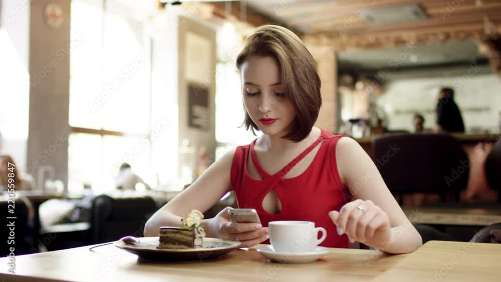 nice girl with short hair and in a red dress sits in a cafe talking on the phone and going to have coffee with dessert nice girl with short hair and in a red dress sits in a cafe talking on the phone and going to have coffee with dessert