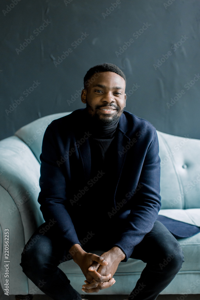 Young black man in coat on dark background sitting on a blue sofa crossed his fingers Young black man in coat on dark background sitting on a blue sofa crossed his fingers
