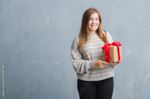 Young adult woman over grey grunge wall holding a present with a happy face standing and smiling with a confident smile showing teeth