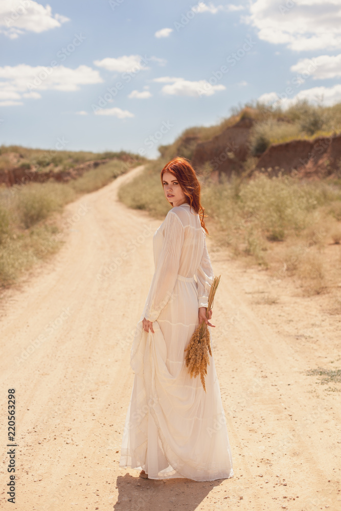 portrait of a beautiful red-haired girl in a white vintage wedding dress in full length with a bouquet of reeds standing with her back on the road and nature background