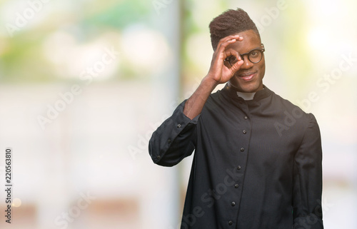 Young african american priest man over isolated background doing ok gesture with hand smiling, eye looking through fingers with happy face.