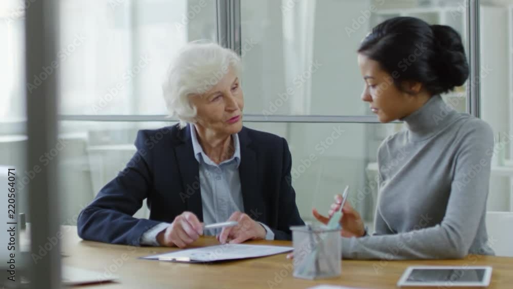 Medium shot of two multiethnic female colleagues sitting at desk, studying business documents and discussing work in office Medium shot of two multiethnic female colleagues sitting at desk, studying business documents and discussing work in office