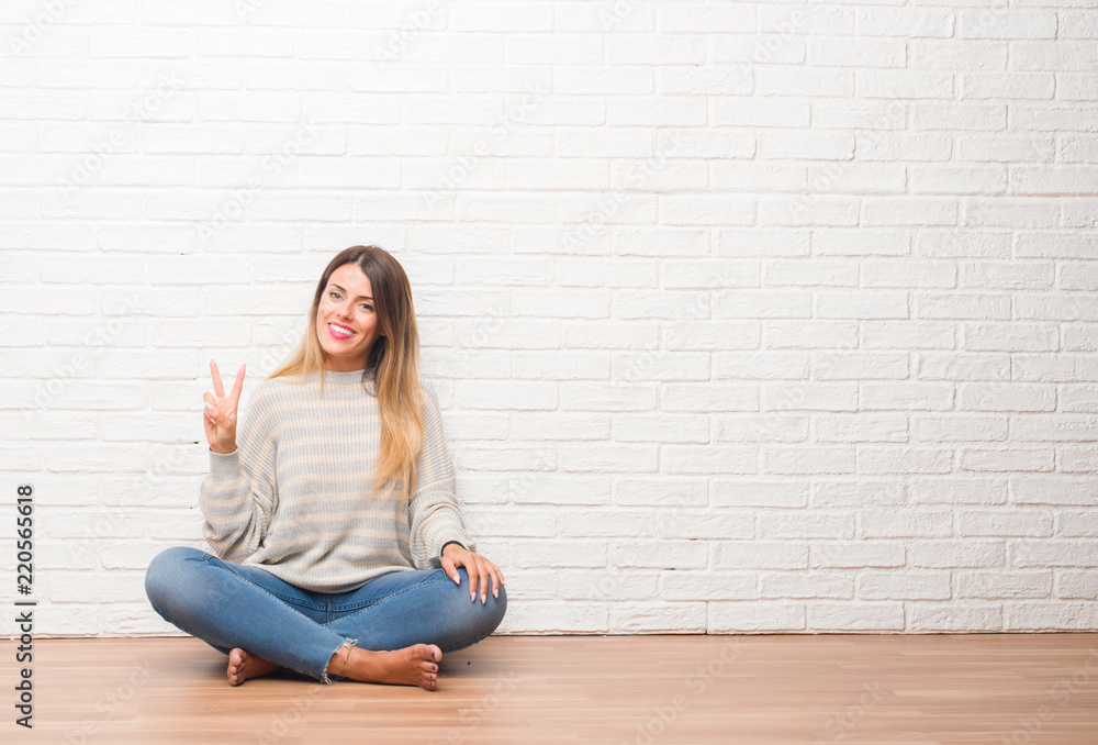 Young adult woman sitting on the floor over white brick wall at home showing and pointing up with fingers number two while smiling confident and happy. Young adult woman sitting on the floor over white brick wall at home showing and pointing up with fingers number two while smiling confident and happy.