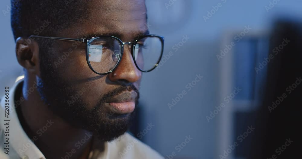 Close up of the bored young African American man in glasses sitting in front of the computer screen and looking sleepy at night in the office. Close up of the bored young African American man in glasses sitting in front of the computer screen and looking sleepy at night in the office.