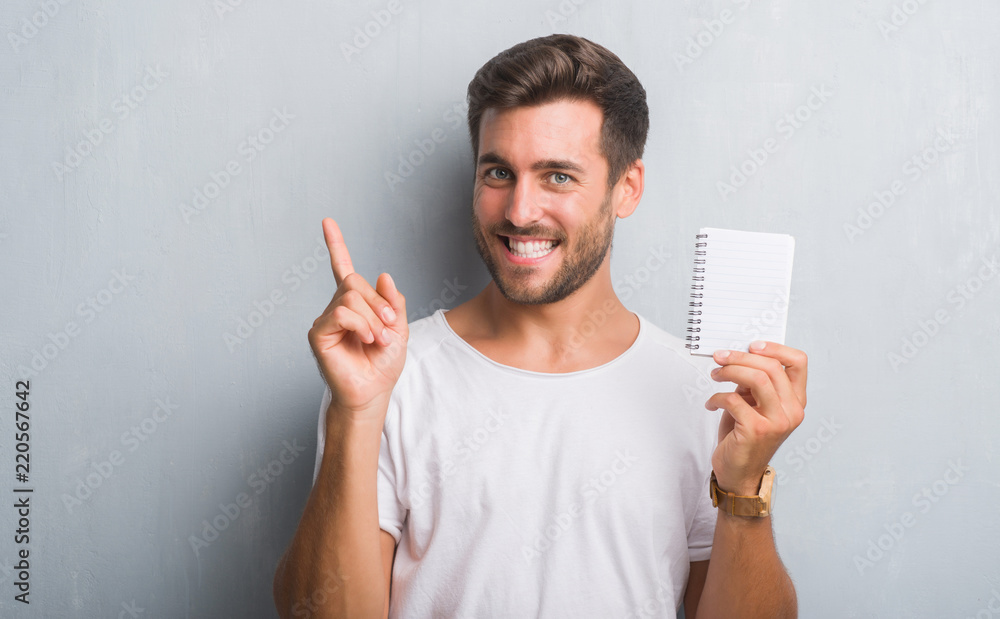 Handsome young man over grey grunge wall holding blank notebook very happy pointing with hand and finger to the side Handsome young man over grey grunge wall holding blank notebook very happy pointing with hand and finger to the side