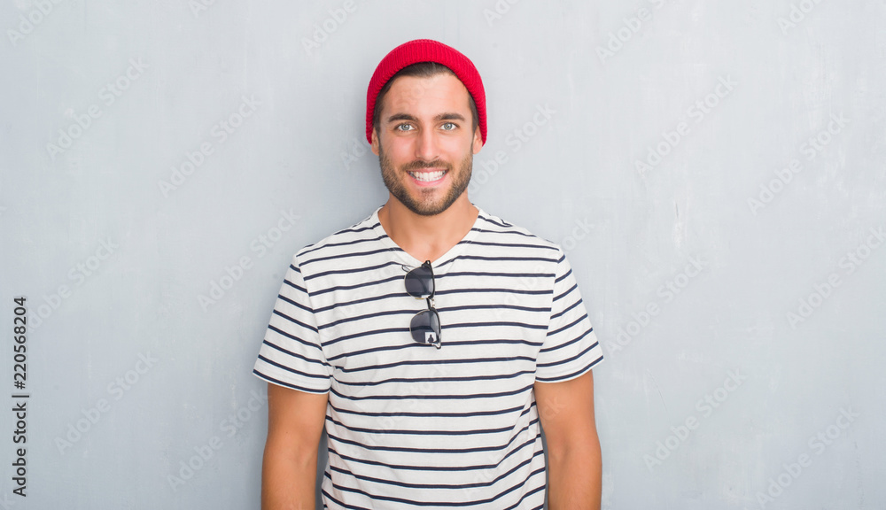 Fototapeta premium Handsome young hipster man over grey grunge wall wearing navy t-shirt and wool cap with a happy and cool smile on face. Lucky person.