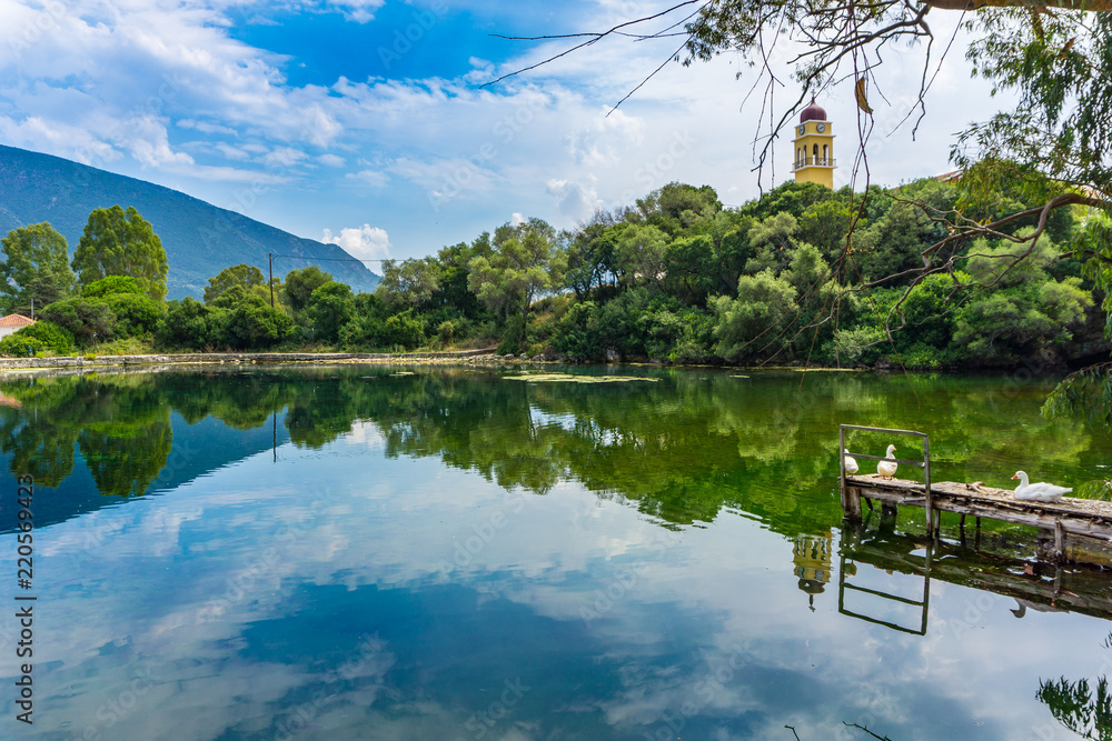 Picturesque view of Karavomilos lake at Sami in Kefalonia ionian island of  Greece. Perfect reflection of the claoudscape, the trees and church on the  calm waters of the lake. Stock Photo