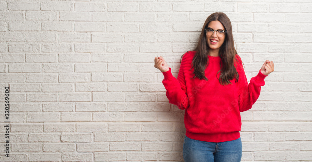 Young brunette woman standing over white brick wall celebrating surprised and amazed for success with arms raised and open eyes. Winner concept.