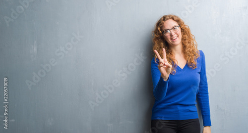 Young redhead woman over grey grunge wall smiling looking to the camera showing fingers doing victory sign. Number two.