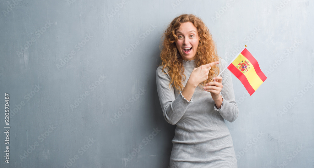 Young redhead woman over grey grunge wall holding flag of Spain very happy pointing with hand and finger Young redhead woman over grey grunge wall holding flag of Spain very happy pointing with hand and finger