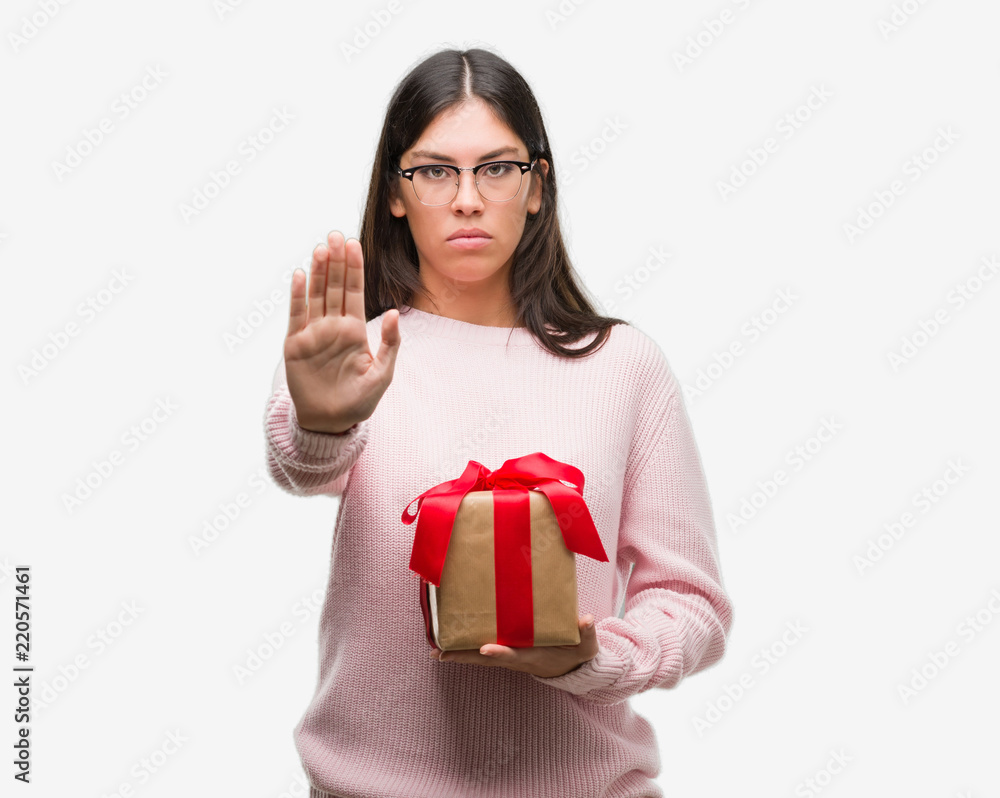 Young hispanic woman holding a gift with open hand doing stop sign with serious and confident expression, defense gesture Young hispanic woman holding a gift with open hand doing stop sign with serious and confident expression, defense gesture