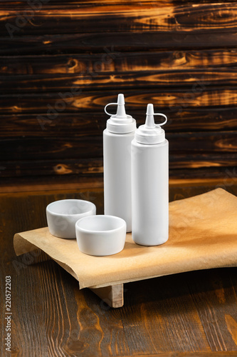 Vertical mockup of white plastic bottles and two sauceboats on a tray at wooden background with free space for branding.