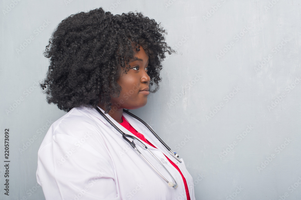 Young african american doctor woman over grey grunge wall looking to side, relax profile pose with natural face with confident smile. Young african american doctor woman over grey grunge wall looking to side, relax profile pose with natural face with confident smile.
