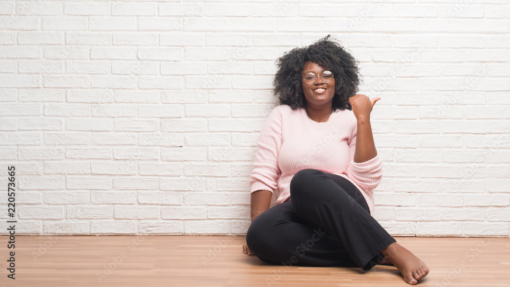 Young african american woman sitting on the floor at home smiling with happy face looking and pointing to the side with thumb up. Young african american woman sitting on the floor at home smiling with happy face looking and pointing to the side with thumb up.