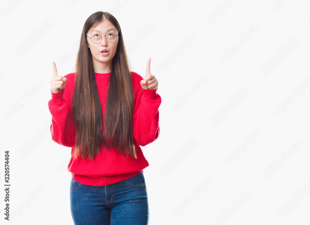 Young Chinese woman over isolated background wearing glasses amazed and surprised looking up and pointing with fingers and raised arms.