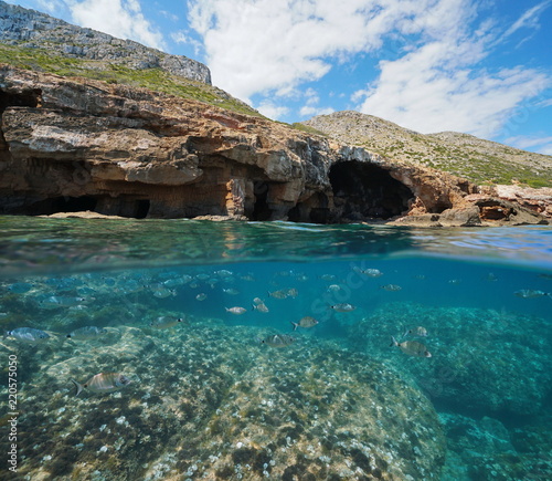 Large cave on the coastline with rocks and a shoal of fish underwater, split view above and below water surface, Mediterranean sea, Cova Tallada, Costa Blanca, Javea, Alicante, Valencia, Spain