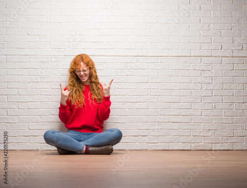 Young redhead woman sitting over brick wall shouting with crazy expression doing rock symbol with hands up. Music star. Heavy concept. © Krakenimages.com