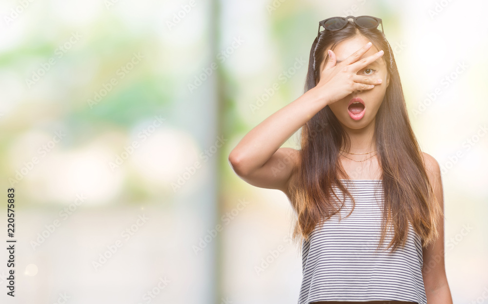 Young asian woman wearing sunglasses over isolated background peeking in shock covering face and eyes with hand, looking through fingers with embarrassed expression.