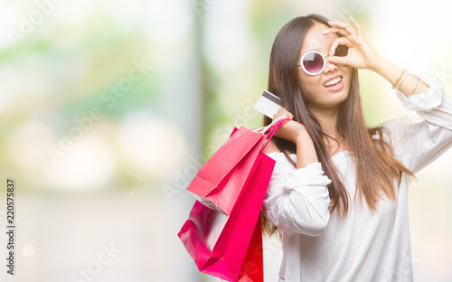 Young asian woman holding shopping bags on sales over isolated background with happy face smiling doing ok sign with hand on eye looking through fingers