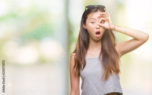 Young asian woman wearing sunglasses over isolated background doing ok gesture shocked with surprised face, eye looking through fingers. Unbelieving expression.