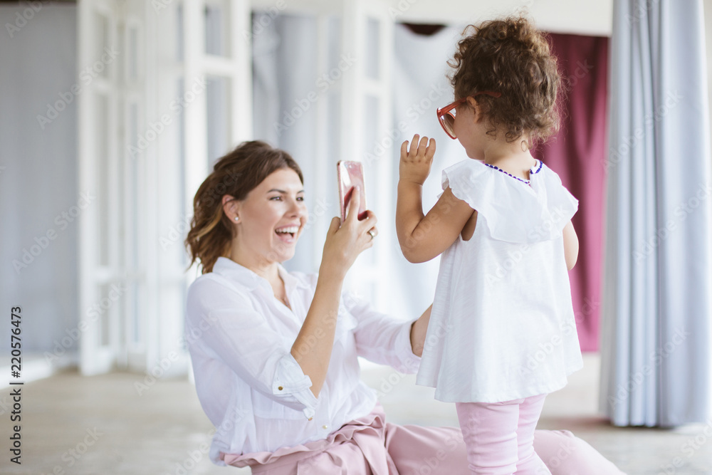 Fototapeta premium Cheerful mother joyfully taking photos on cellphone of pretty little daughter wearing adult sunglasses while spending time together at home