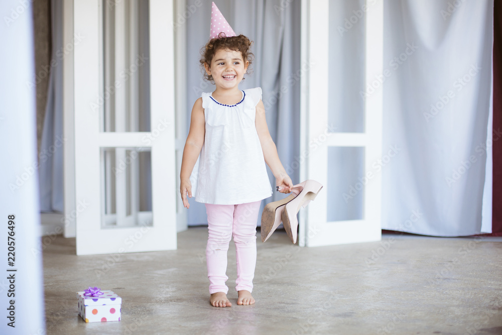 Cheerful little girl with dark curly hair in birthday cap happily looking aside while holding shoes on heels in hand with gift box near on floor at home
