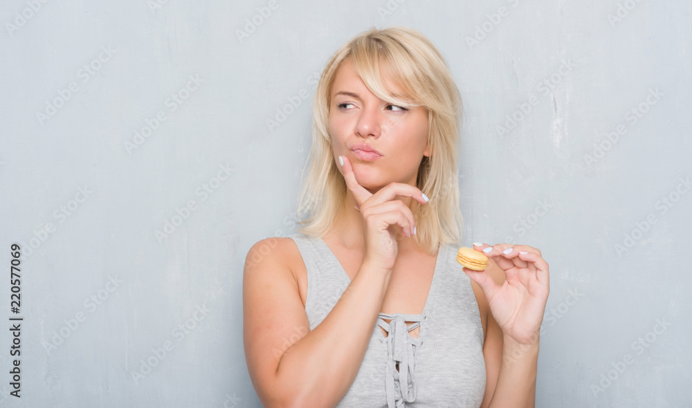 Adult caucasian woman over grunge grey wall eating macaron serious face thinking about question, very confused idea Adult caucasian woman over grunge grey wall eating macaron serious face thinking about question, very confused idea
