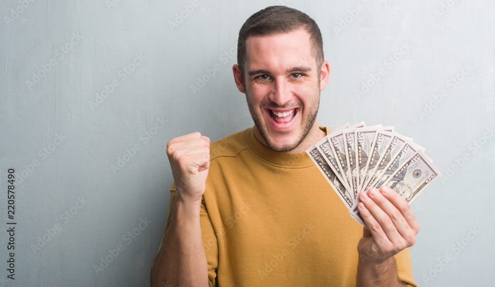 Young caucasian man over grey grunge wall holding dollars screaming proud and celebrating victory and success very excited, cheering emotion Young caucasian man over grey grunge wall holding dollars screaming proud and celebrating victory and success very excited, cheering emotion