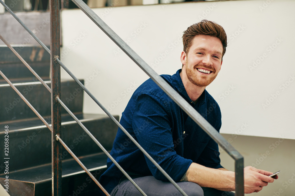 Smiling young businessman sitting on office stairs reading text Smiling young businessman sitting on office stairs reading text
