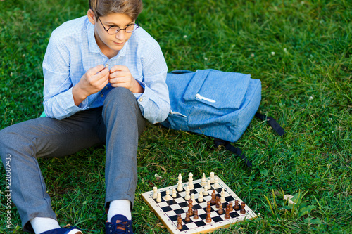 cute boy in riound glasses and blue shirt sits on the grass in the park and plays chess at wooden chessboard. Hobby, education, intellectual game concept photo