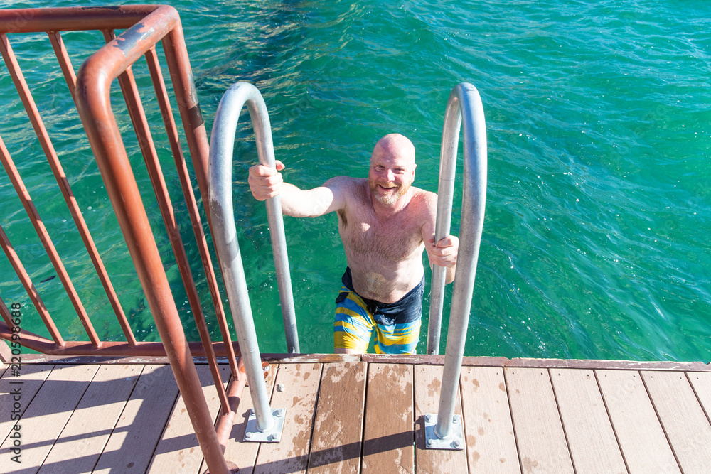 Fototapeta premium Shirtless, bald man climbing out of lake on a ladder