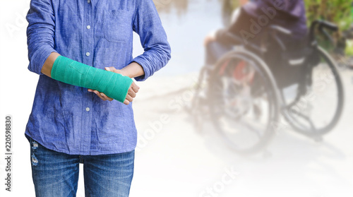 broken arm, injury woman standing wearing shirt and jeans with green cast on hand isolated on blurred background patient sitting on wheelchair
