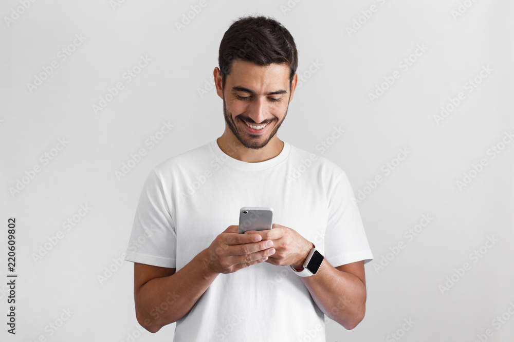 Daylight photo of young european man pictured isolated on grey background dressed in casual blank t-shirt holding smartphone with bond hands and smiling positively while looking at it Daylight photo of young european man pictured isolated on grey background dressed in casual blank t-shirt holding smartphone with bond hands and smiling positively while looking at it