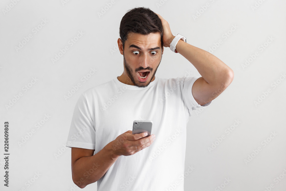 Daylight portrait of handsome surprised young man in white t-shirt, holding smartphone, looking scared and confused. Shock content Daylight portrait of handsome surprised young man in white t-shirt, holding smartphone, looking scared and confused. Shock content