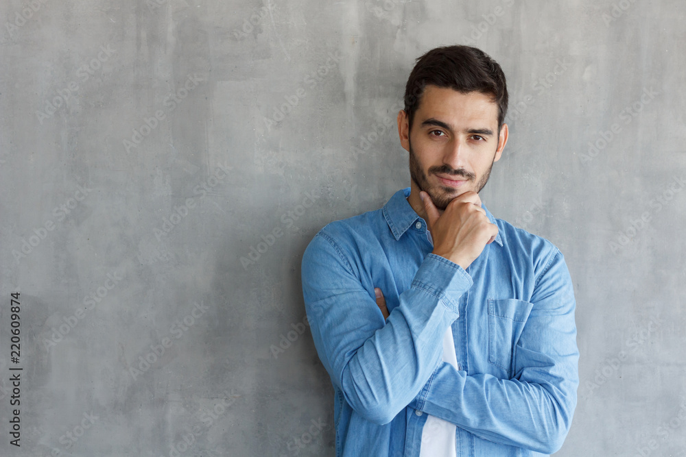 Portrait of thoughtful man in blue shirt, touching his chin, standing against gray textured wall with copy space for your ads Portrait of thoughtful man in blue shirt, touching his chin, standing against gray textured wall with copy space for your ads