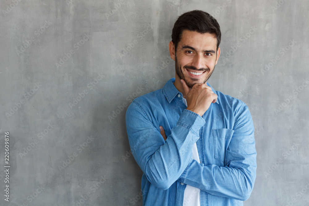Portrait of smiling handsome man in blue shirt, touching his chin, standing against gray textured wall with copy space for your ads Portrait of smiling handsome man in blue shirt, touching his chin, standing against gray textured wall with copy space for your ads