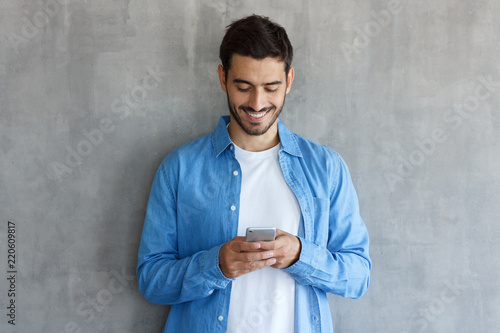 Young man standing against gray textured wall, looking at screen of smartphone, browsing web and smiling nicely while chatting