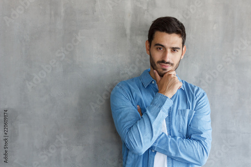 Portrait of thoughtful man in blue shirt, touching his chin, standing against gray textured wall with copy space for your ads