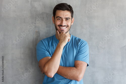 Portrait of smiling man in blue t-shirt, touching his chin, standing against gray textured wall
