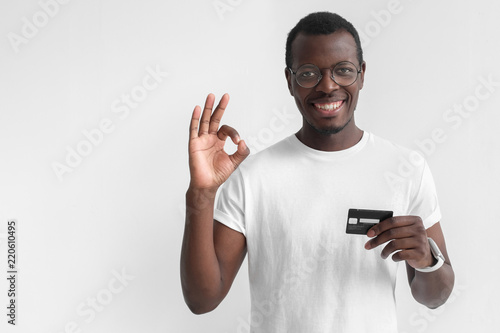 Young smiling dark skin african man in white t shirt holding credit card and showing okay sign isolated on gray background with copy space