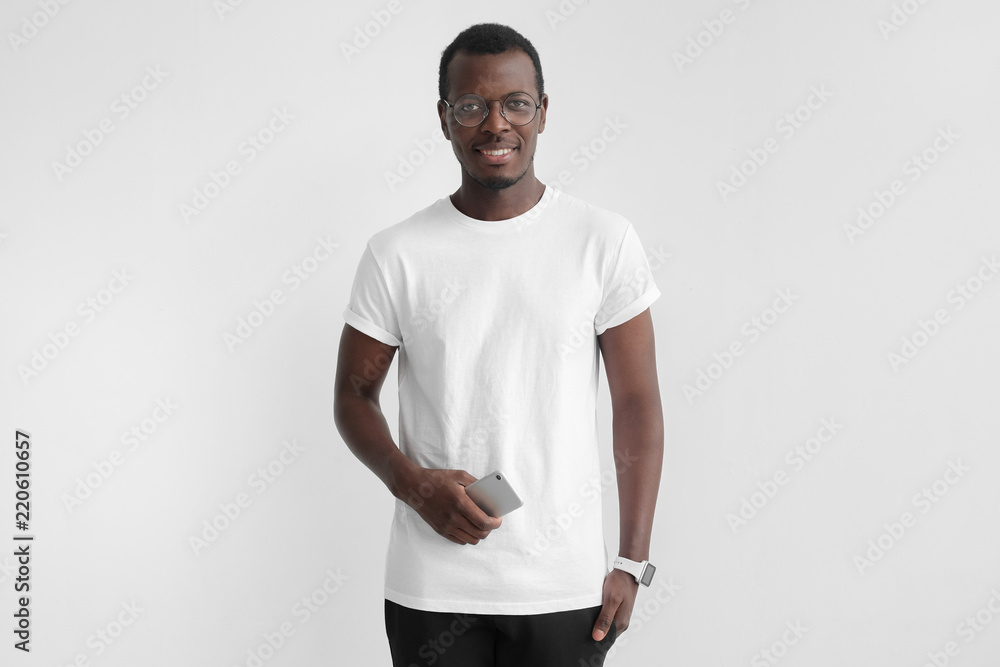 Young african american man in white t-shirt and spectacles isolated on gray background, holding cellphone in hand, looking at camera with open smile Young african american man in white t-shirt and spectacles isolated on gray background, holding cellphone in hand, looking at camera with open smile