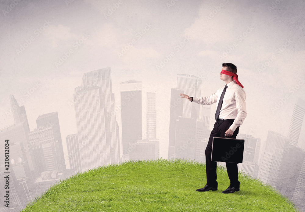 Young blindfolded businessman steps on a a patch of grass with a cloudy city in the background Young blindfolded businessman steps on a a patch of grass with a cloudy city in the background